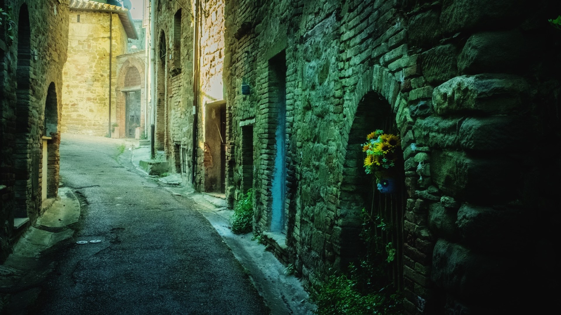 A narrow stone street curving towards the light, with flowers beside a doorway